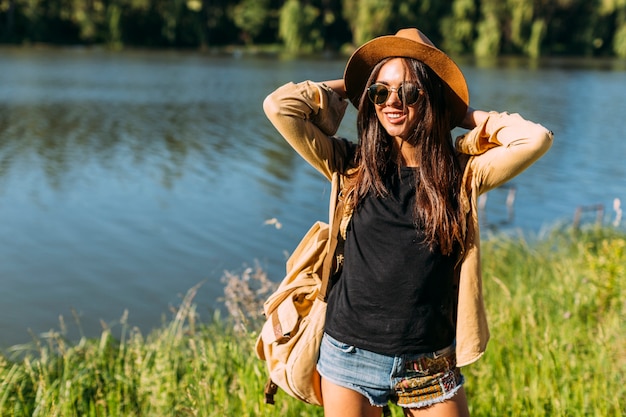 Free Photo young female traveler with backpack, glasses and hat standing in front of lake