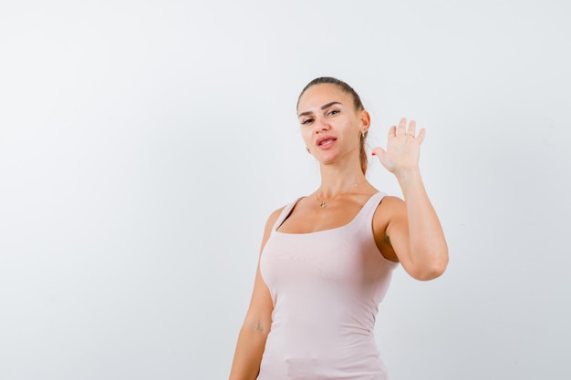 Young female waving hand for greeting in white tank top and looking happy , front view.