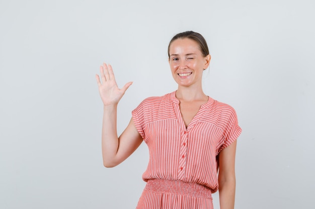 Young female waving hand and winking eye in striped dress and looking cheery , front view.