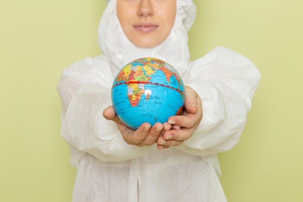 young female in white special suit and yellow protective helmet holding little round globe with slight smile on green