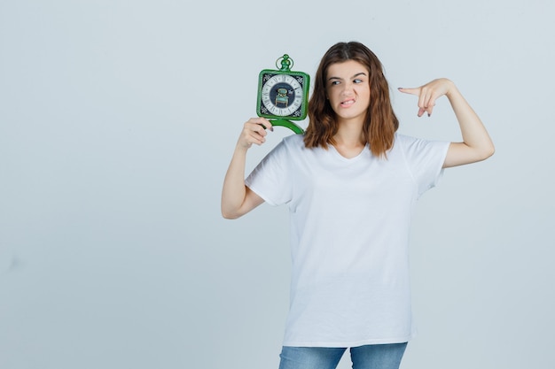 Young female in white t-shirt, jeans pointing at clock, frowning face and looking troubled , front view.