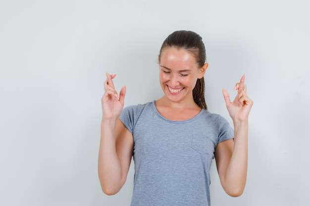 Young female wishing good luck with crossed fingers in grey t-shirt and looking happy , front view.