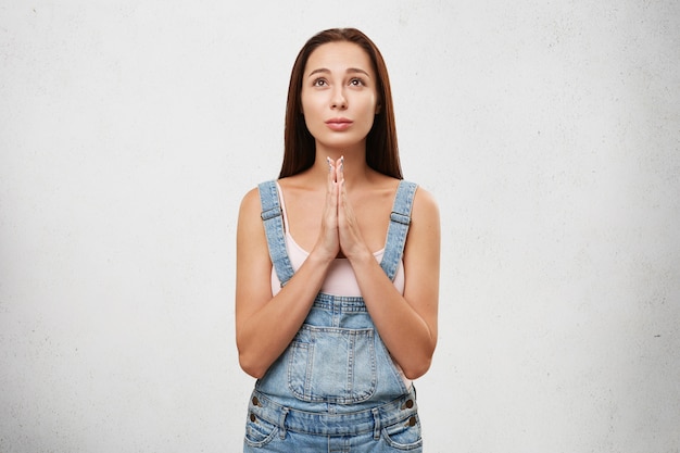 Young female with long dark straight hair looking up hopefully praying with her hands folded