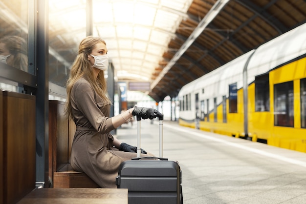 Free photo young female with a suitcase wearing a facemask and gloves and waiting at a train station