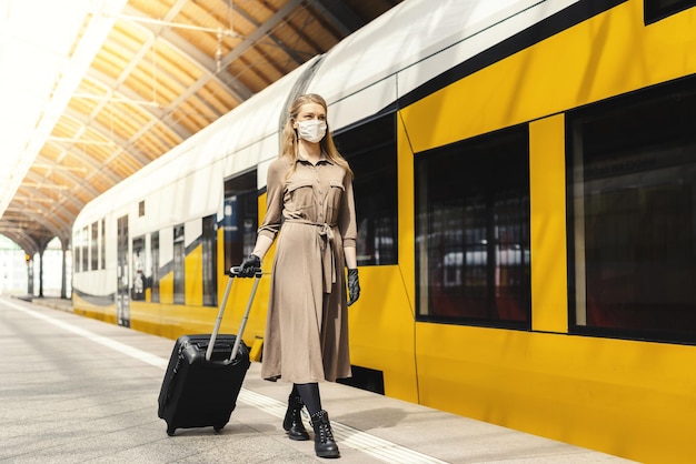 Young female with a suitcase wearing gloves and a facemask walking at a train station - COVID-19