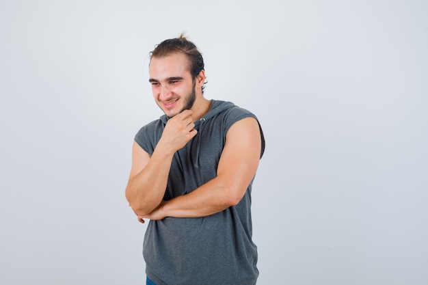 Young fit male keeping hand under chin in sleeveless hoodie  and looking blissful , front view.