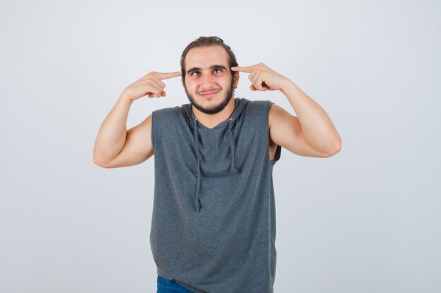 Young fit male pointing at  squint eyes in sleeveless hoodie  and looking merry , front view.