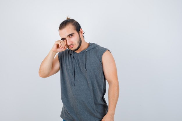 Young fit male rubbing his eye in sleeveless vest and looking upset , front view.