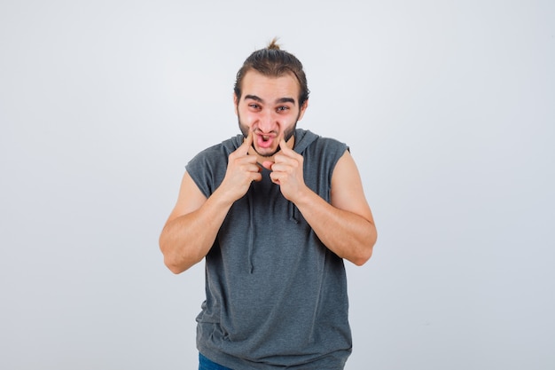 Young fit male in sleeveless hoodie 
 pressing cheeks with fingers and looking glad , front view.