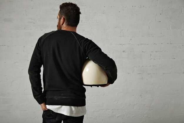 Young fit motorcyclist in black sweatshirt and jeans holding a white helmet on his side with painted brick walls