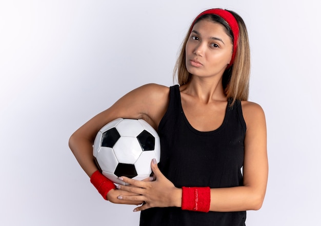 Young fitness girl in black sportswear and red headband holding soccer ball  with serious face standing over white wall