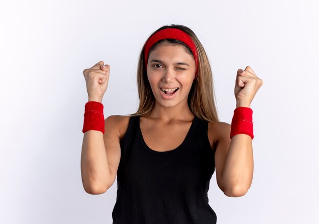 Young fitness girl in black sportswear and red headband lookign  clenching fists winking and smiling standing over white wall