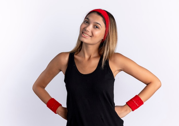 Free photo young fitness girl in black sportswear and red headband  smiling cheerfully with arms at hip standing over white wall