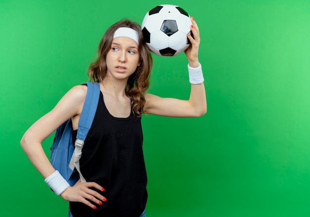 Free Photo young fitness girl in black sportswear with backpack and headband holding soccer over head lookign aside puzzled standing over green wall