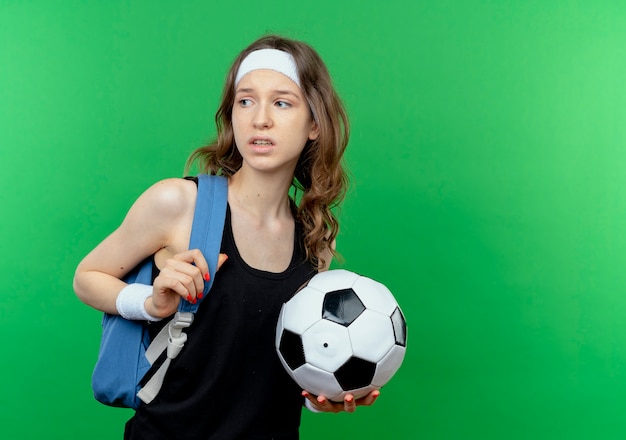 Young fitness girl in black sportswear with backpack and headband holding soccer looking aside confused standing over green wall