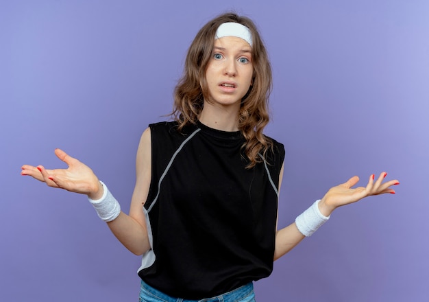 Young fitness girl in black sportswear with headband confused spreading arms to the sides over blue