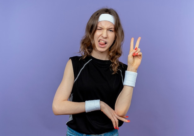 Young fitness girl in black sportswear with headband  having fun sticking out tongue showing victory sign standing over blue wall