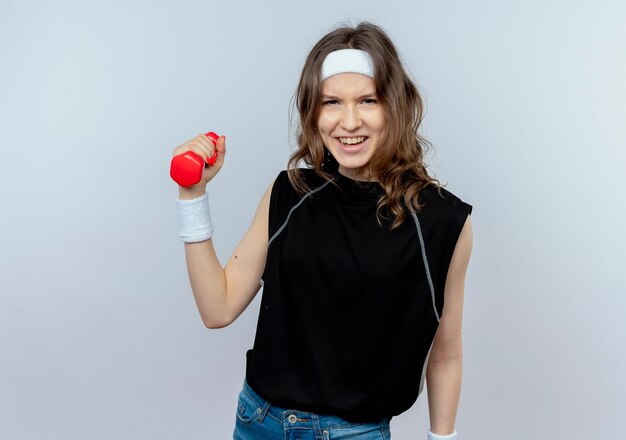 Young fitness girl in black sportswear with headband holding dumbbell in raised hand looking confident standing over white wall