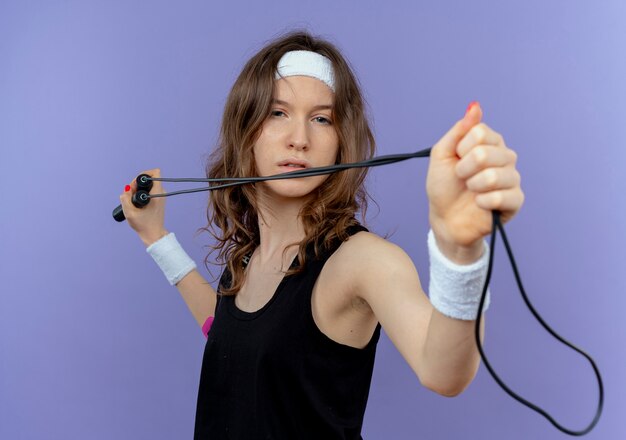 Young fitness girl in black sportswear with headband holding skipping rope like aiming with arrow and bow standing over blue wall