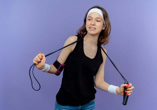 Young fitness girl in black sportswear with headband holding skipping rope lookign confident standing over blue wall