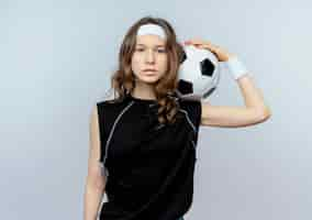 Free photo young fitness girl in black sportswear with headband holding soccer ball  with serious face standing over white wall