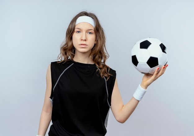 Young fitness girl in black sportswear with headband holding soccer ball  with serious face standing over white wall