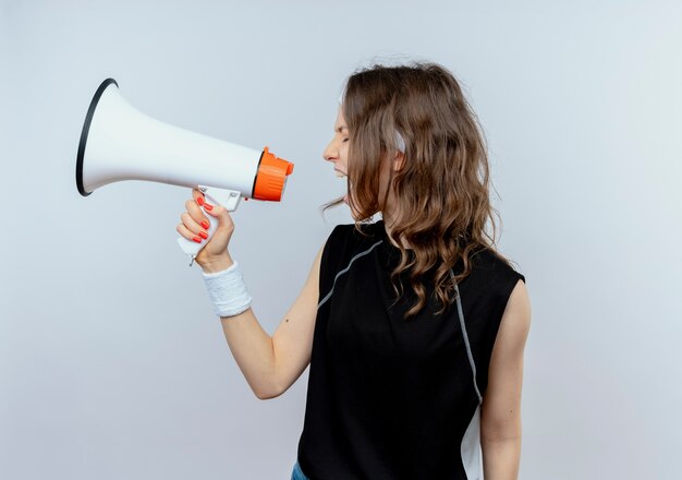 Young fitness girl in black sportswear with headband shouting to megaphone standing over white wall