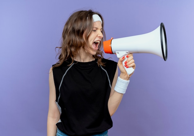 Young fitness girl in black sportswear with headband shouting to megaphone with aggressive expression standing over blue wall