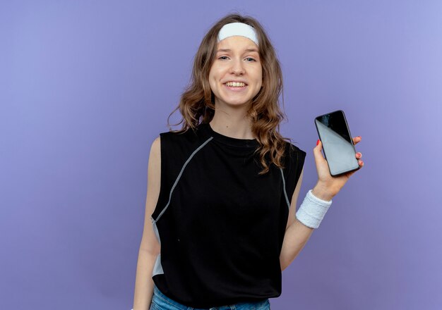 Young fitness girl in black sportswear with headband showing smartphone smiling cheerfully standing over blue wall