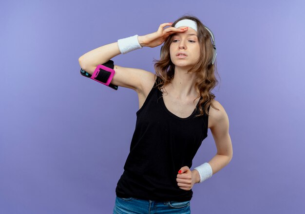 Young fitness girl in black sportswear with headband and smartphone armband looking far away with hand over head standing over blue wall