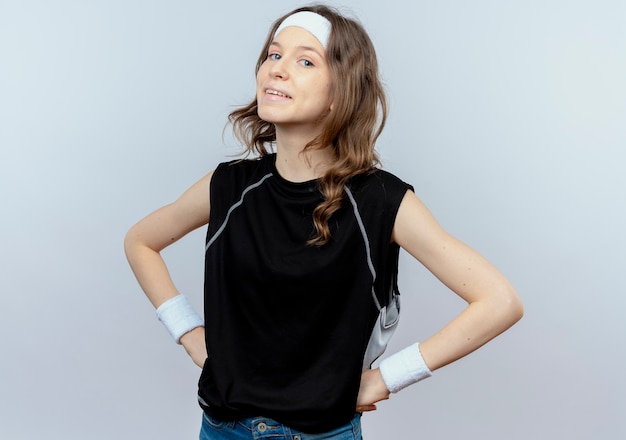 Free photo young fitness girl in black sportswear with headband  smiling confident with arms at hip standing over white wall