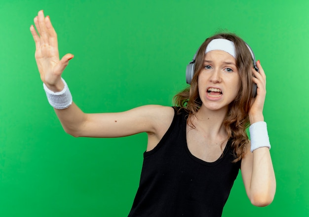 Free photo young fitness girl in black sportswear with headband with headphones looking confused and displeased making stop sign with hand standing over green wall