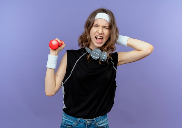 Young fitness girl in black sportswear with headband working out with dumbbell looking confused over blue