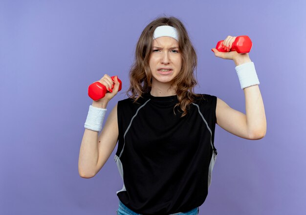 Young fitness girl in black sportswear with headband working out with dumbbells looking confused standing over blue wall