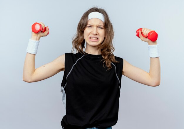 Young fitness girl in black sportswear with headband working out with dumbbells looking strained standing over white wall