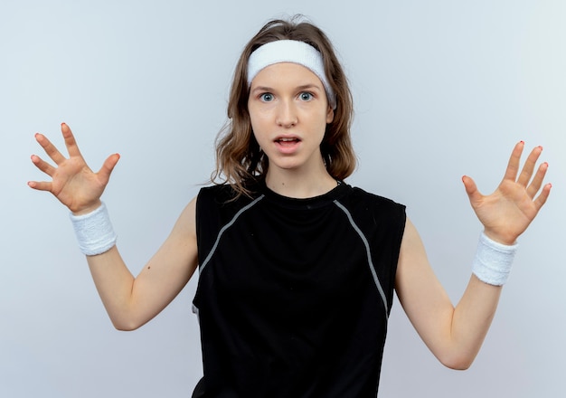 Free photo young fitness girl in black sportswear with headband  worried with raised arms standing over white wall