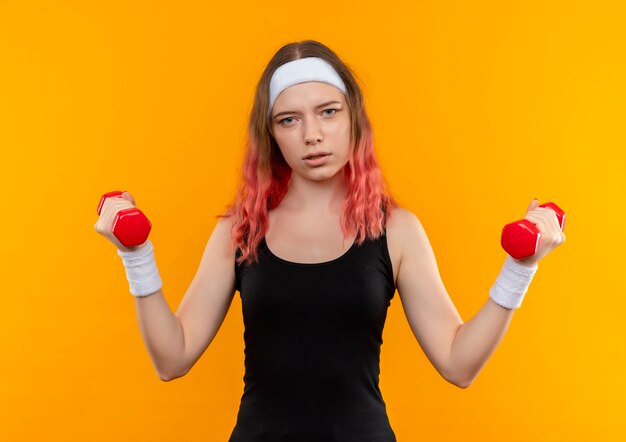 Young fitness girl in sportswear using dumbbells doing exercises looking confident standing over orange wall