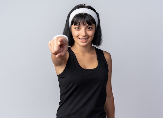 Young fitness girl wearing headband happy and confident pointing with index finger at camera standing over white background