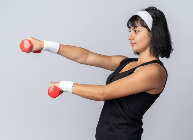 Young fitness girl wearing headband holding dumbbells doing exercises looking confident standing over white background