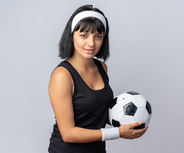 Young fitness girl wearing headband holding soccer ball looking at camera smiling confident standing over white