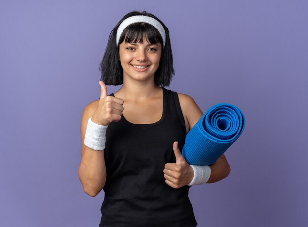 Young fitness girl wearing headband holding yoga mat looking at camera smiling cheerfully showing thumbs up standing over blue background