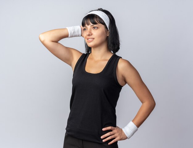 Young fitness girl wearing headband looking aside smiling holding hand on her head thinking positive standing over white