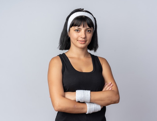 Free photo young fitness girl wearing headband looking at camera with serious confident expression with arms crossed standing over white