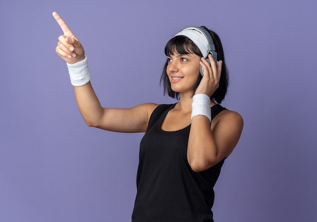 Free photo young fitness girl wearing headband with headphones looking aside with smile on face pointing with index finger at something