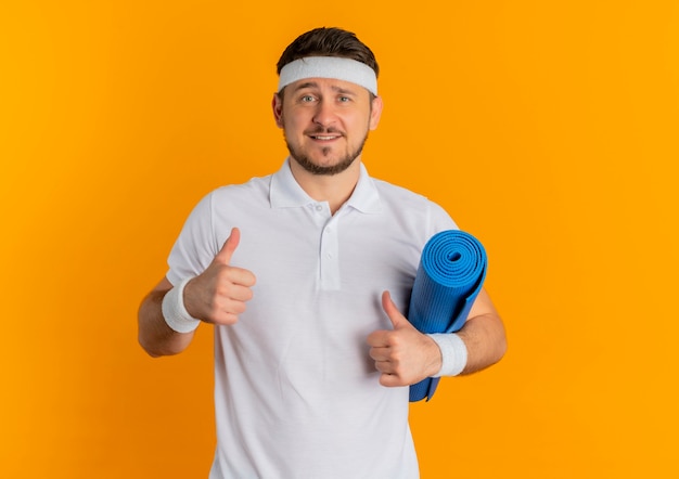 Young fitness man in white shirt with headband holding yoga mat showing thumbs up looking at camera smiling standing over orange background