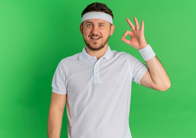 Young fitness man in white shirt with headband looking to the front smiling cheerfully doing ok sing standing over green wall