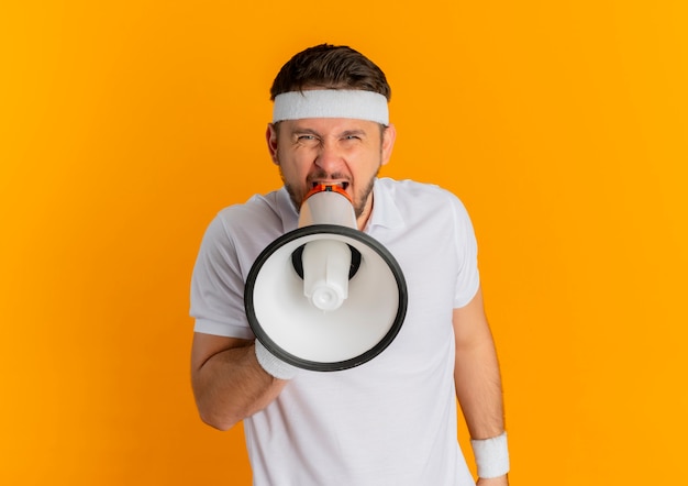 Free photo young fitness man in white shirt with headband shouting to megaphone with aggressive expression standing over orange wall