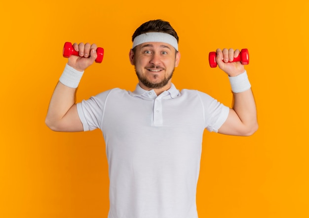 Young fitness man in white shirt with headband working out with dumbbells looking confident smiling standing over orange wall