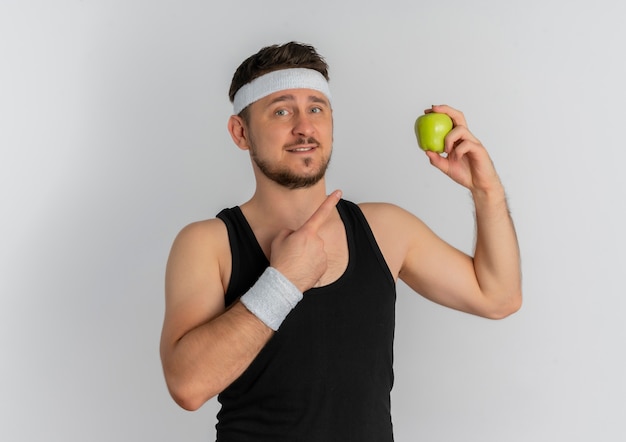 Young fitness man with headband holding green apple pointing with finger to it smiling cheerfully standing over white background