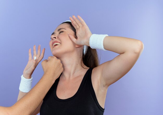 Young fitness woman in headband being punched with fist in her face standing over blue background